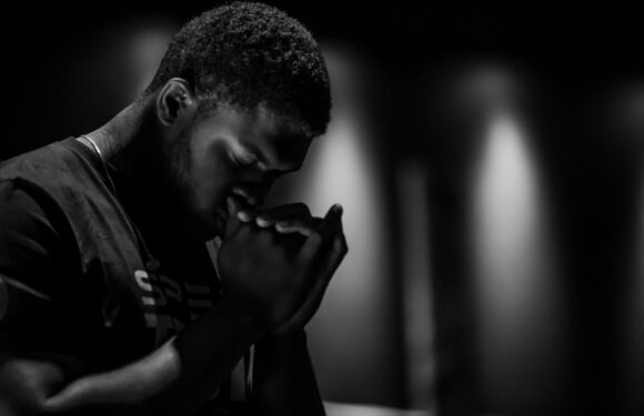 A young man in a dark Pizza Ranch uniform prays with hands clasped, head bowed and eyes closed in a dimly lit setting.