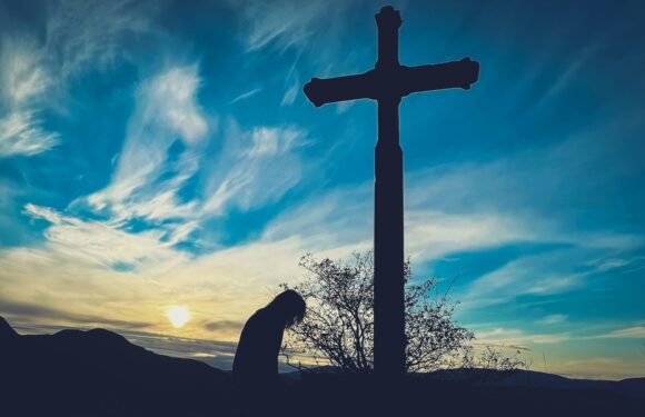 A person kneels in prayer beside a large cross on a hillside during sunset, with a sky filled with clouds and a silhouette of mountains in the background.