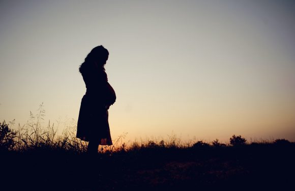 A pregnant woman stands outdoors on a hill during sunset, holding her belly and looking down contemplatively.