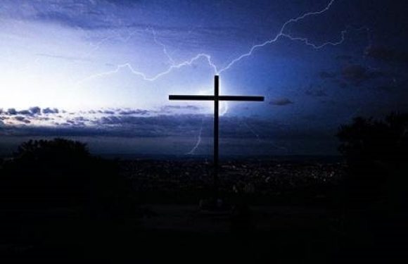 A cross on a hilltop illuminated by lightning in a dark, stormy sky with clouds and distant city lights in the background.