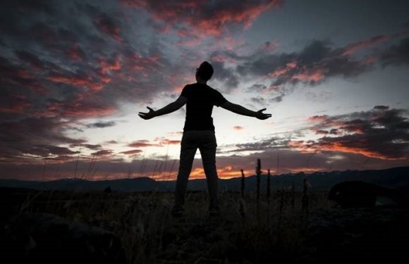 A person stands outdoors at sunset with arms outstretched, silhouetted against a colorful sky with clouds, mountains, and cacti in the background.