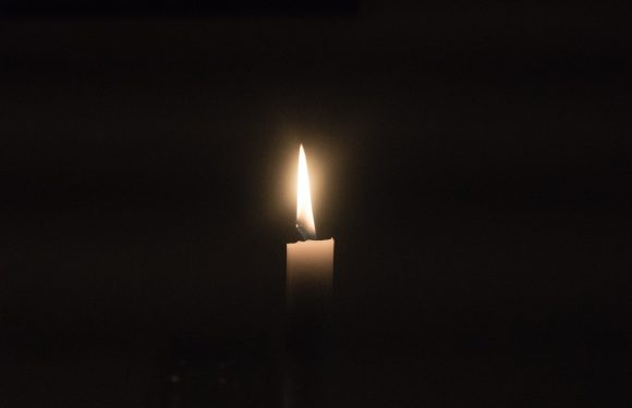 A single candle flame flickers in a dark background, casting a soft glow around the small, white candle.