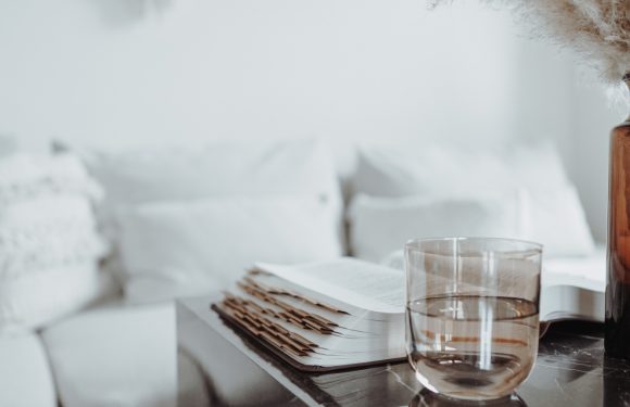 An open book and a glass of water sit on a black marble table, with a white couch and decorative wall hanging in a bright, minimal living room.