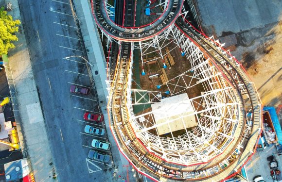 An aerial view of a roller coaster under construction at an amusement park, with some parked cars and pathways nearby.