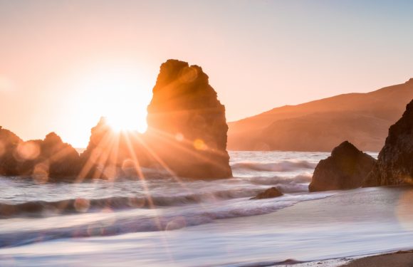 Sunset over a rocky coastline with the sun partially obscured by a large rock formation, casting warm light and lens flare over the waves and sandy shore.