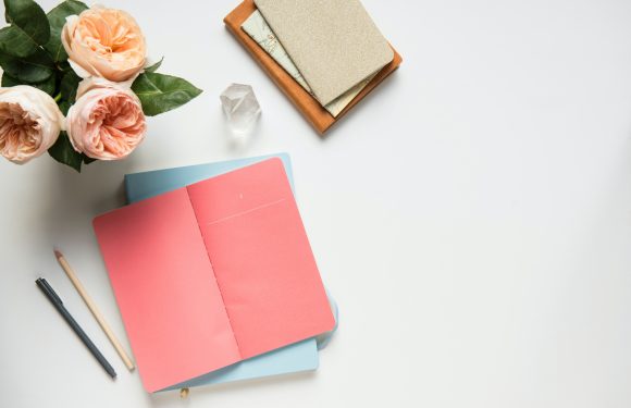 A top-down view of a white desk with pink roses, an open pink and blue notebook, two pens, a small stack of notebooks, and a crystal on it.