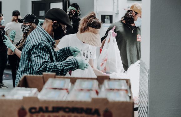 People wearing masks and gloves gather around food packages at a distribution event, with some holding bags and others working with food boxes.