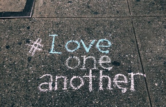 Pink and blue chalk writing on a concrete sidewalk reads "#Love one another."