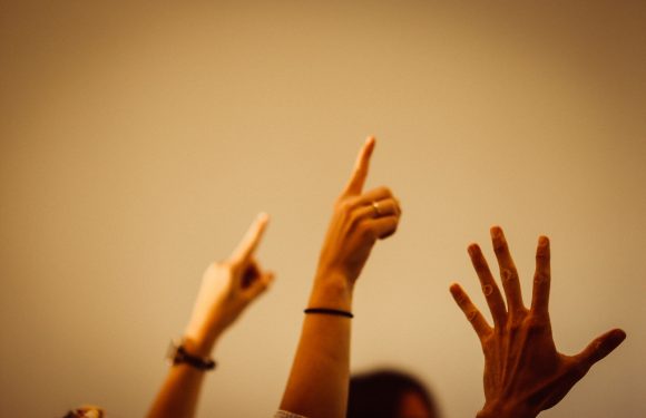Three hands are raised against a plain beige background, each with fingers extended or pointing, some adorned with rings and bracelets.