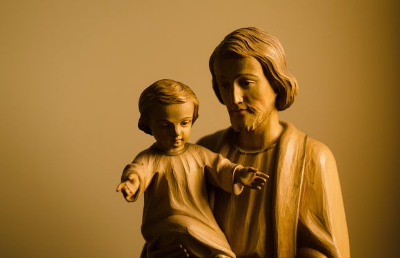 A wooden sculpture of a man holding a child, both with detailed facial features and flowing hair, set against a plain, neutral background.