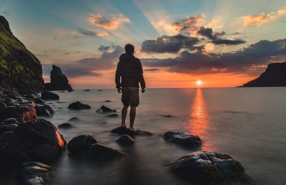 A person stands barefoot on rocks in the water, facing a sunset over the ocean with clouds and distant cliffs on both sides.