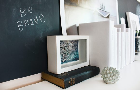 A white picture frame, a black and gold book, a small sculpture resembling a pine cone, and a row of white rolled papers are placed on a white surface against a blackboard with "Be Brave" written in chalk.