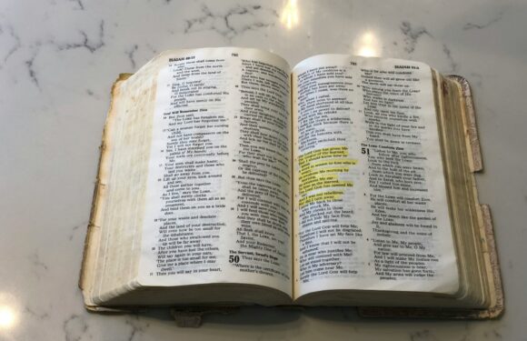 An open Bible resting on a white, reflective surface with sunlight glinting off of it; some text is highlighted yellow, with the highlighted portion on the right page.