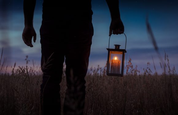 A person dressed in dark clothing holding a lantern with a candle inside, standing in a field at dusk or night with a sunset or twilight sky.