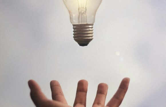 A hand appears to be tossing a lit lightbulb into the air against a cloudy sky background.