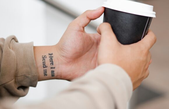 A person in a beige jacket holds a black coffee cup with a white lid, showing a tattoo that says "Here I am Send me" on their wrist.