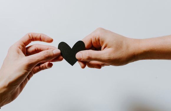 Two hands are exchanging a black heart-shaped cutout against a plain background, symbolizing sharing love or kindness.