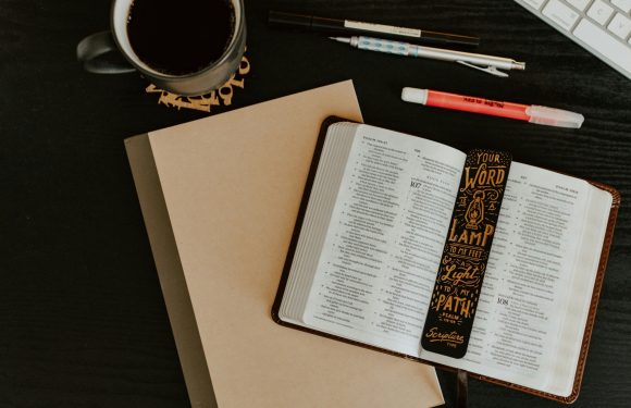 Open Bible with a decorative book marker, a closed beige notebook, a cup of coffee, pens, and a keyboard on a dark wooden desk.