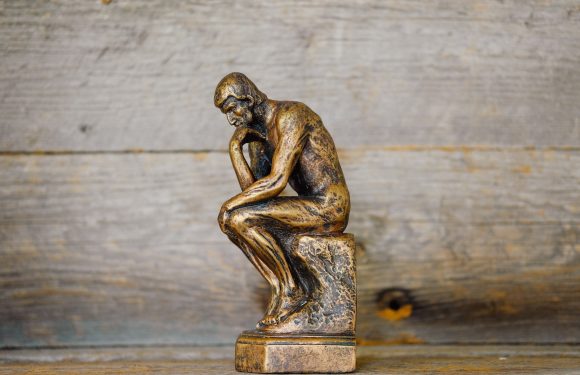 A small bronze-colored sculpture of a man sitting on a block with his chin resting on his hand, appearing deep in thought, placed on a wooden surface.