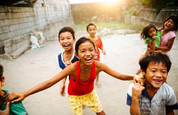 Children laughing and playing outdoors on a dusty ground, with a brick wall and wooden fence in the background, sunlight glowing in the late afternoon.