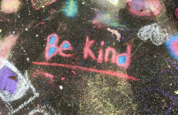 Colorful chalk drawings and doodles on a black pavement surface, including the phrase "be kind" written in pink with a red underline.