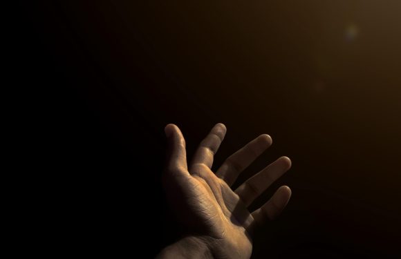 A hand reaching upward in a dimly lit environment with warm light illuminating the fingers, against a mostly dark background with some soft light spots.