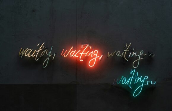 Neon signs spell out "waiting" repeatedly in red, yellow, and turquoise, with the central "waiting" illuminated in red, against a dark wall background.