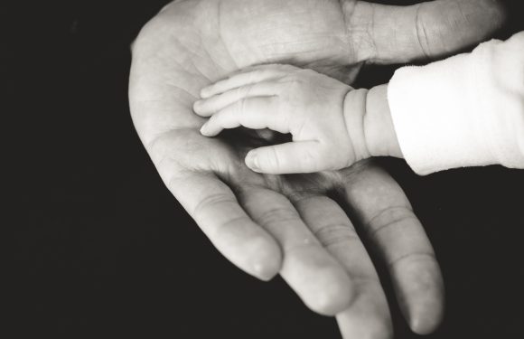 A tiny baby hand touches an adult hand, creating a sense of connection and tenderness. The image is in black and white with a dark background.