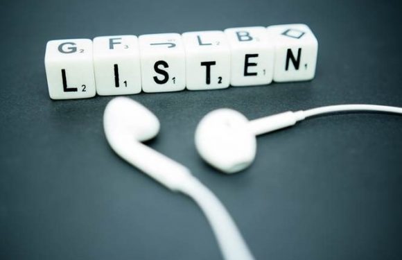 Scrabble tiles spell out "LISTEN" with a pair of white earbud headphones below on a dark surface.