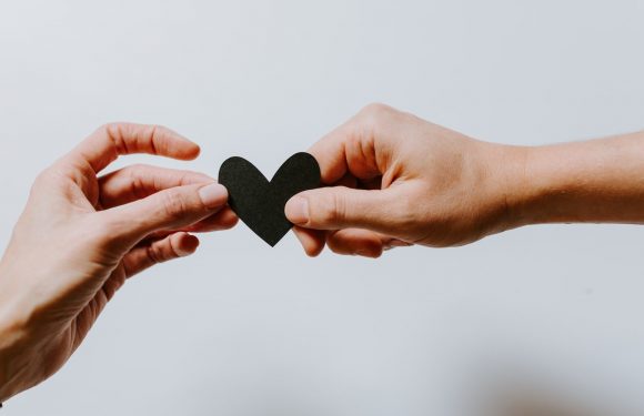 Two hands are exchanging a black heart-shaped object against a plain background. One hand is holding the heart, while the other is reaching to take it.