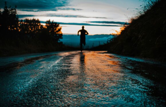 A person running on a wet, reflective road at sunset, with silhouettes of trees on both sides and a sky with layered clouds in the background.