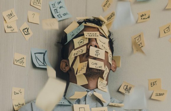 A person with a cluttered face covered in sticky notes, wearing a collared shirt, surrounded by more sticky notes with to-do lists and reminders on a wall.
