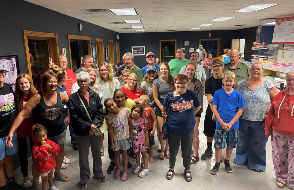 Group of people, including children and adults, gathered inside a restaurant with dark walls, tile flooring, and wooden trim around the windows.