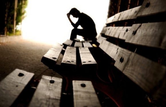 A person sitting on a park bench with their head resting in one hand, appearing to be in deep thought, in an outdoor setting with a bright background.