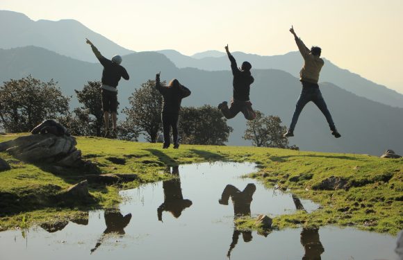 Four people silhouetted, some mid-jump, on a grassy hill near a water reflection with mountains and trees in the background.