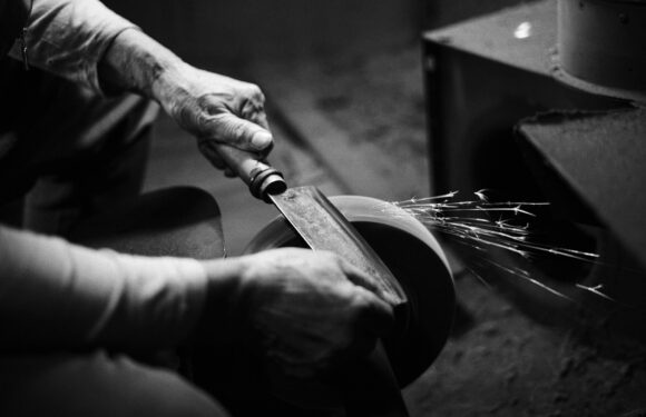 Black and white image of a person using a knife to grind or sharpen a circular metal object, with sparks flying from the contact.