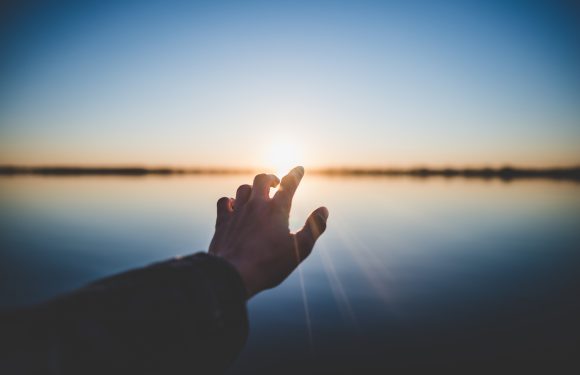 Hand reaching out over a body of water at sunset, with the sun just above the horizon and sky reflected in the water, creating a peaceful scene.