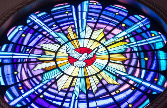 A colorful stained glass window features a central white dove with outstretched wings against a red background, surrounded by radiating blue, yellow, and purple patterns with stars.
