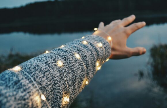 A person's arm wearing a patterned sweater with string lights wrapped around it, reaching out over a calm body of water with a blurred natural landscape in the background.