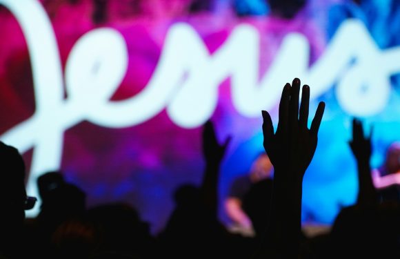 Silhouettes of people raising their hands in front of a colorful, blurred stage with a bright white "Love" sign, creating a lively atmosphere.