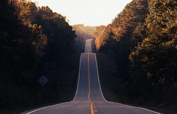 A long, winding road with a double yellow line stretches through a wooded area with fall foliage, ascending into the distance under a bright, clear sky.