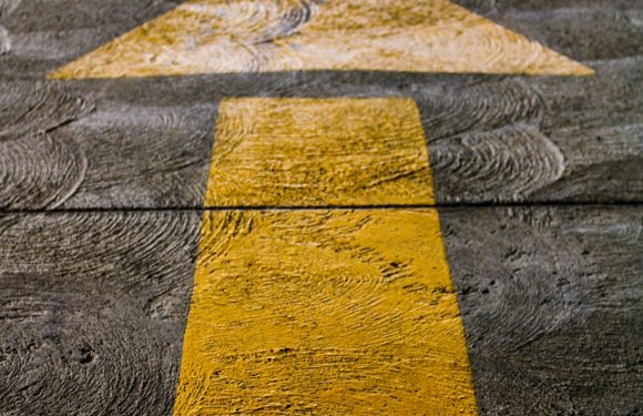 A yellow painted arrow on a wooden floor, pointing forward. The floor has a textured, weathered appearance with visible wood grain.