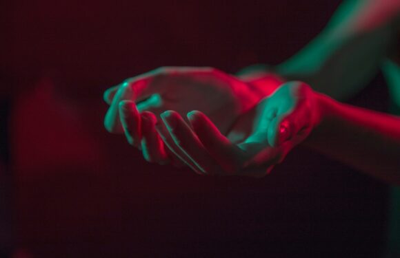 A person's hands are illuminated by red and teal lighting against a black background, with fingers gently curved and palms facing upward.