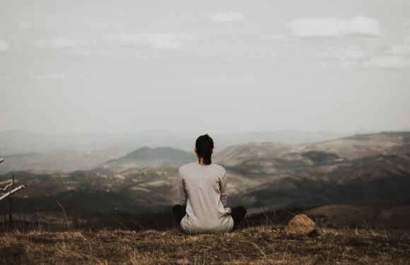 A person sits cross-legged on a grassy hill, facing away, overlooking rolling hills and a cloudy sky in the distance.