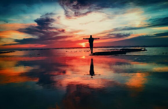 Person standing on rocks near the water with arms outstretched during a colorful sunset or sunrise over the ocean, creating a mirror reflection on the wet surface.
