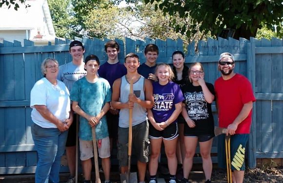A group of nine people, including children and adults, standing outdoors in front of a wooden fence, some holding shovels and a spear, with trees and a house in the background.