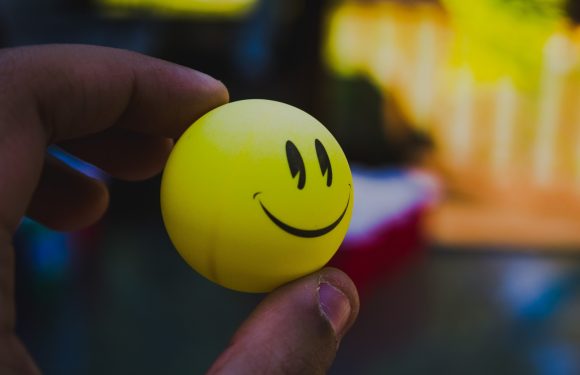 A hand holding a yellow bouncing ball with a black smiley face, partially transparent, with a blurred colorful background.