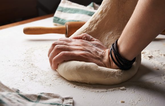 A person kneading dough on a floured surface with a rolling pin and a towel nearby, wearing black bracelets on one wrist.