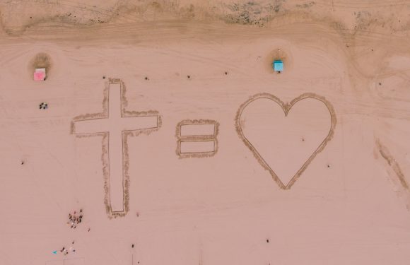 An aerial view of a sandy area with a cross, an equal sign, and a heart drawn in the sand. There are small groups of people and colorful structures nearby.