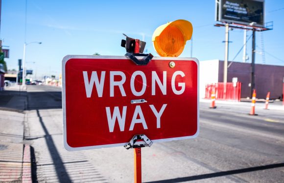A red "Wrong Way" sign with a warning light on top, standing on a street with construction cones and a billboard in the background.